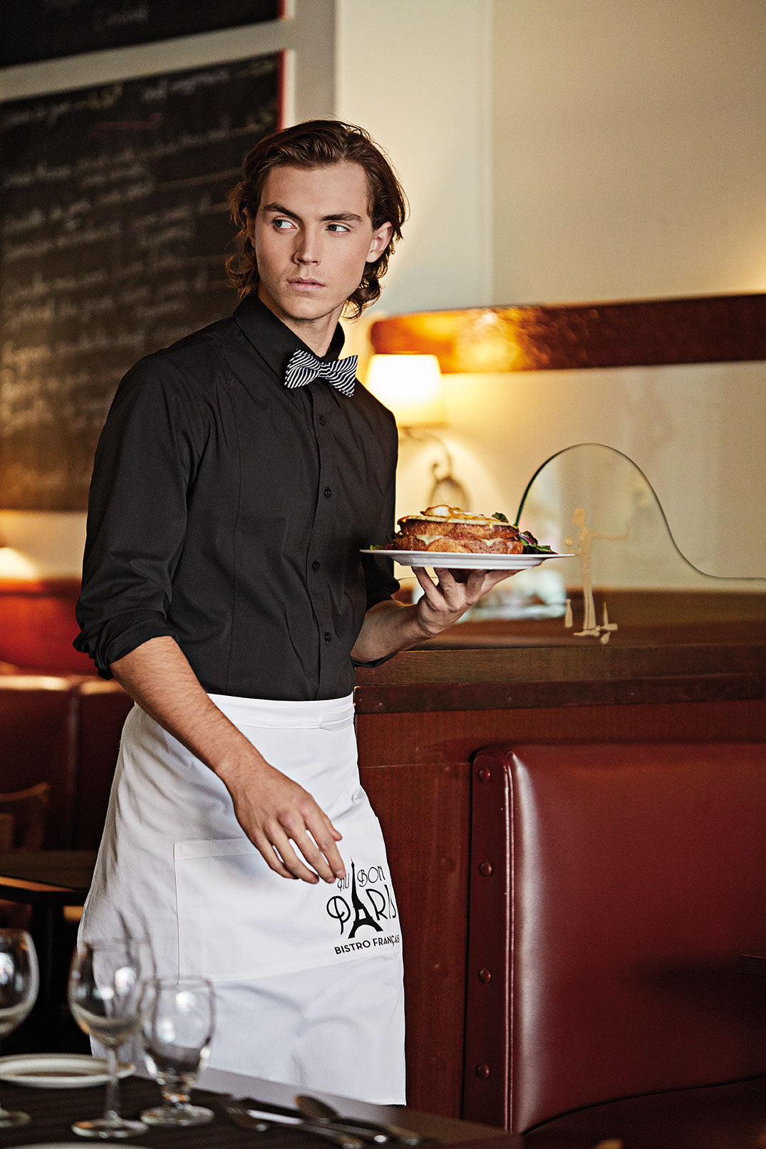 Serveur dans un restaurant qui porte un uniforme composé d’une chemise noire et d’un tablier blanc à la taille